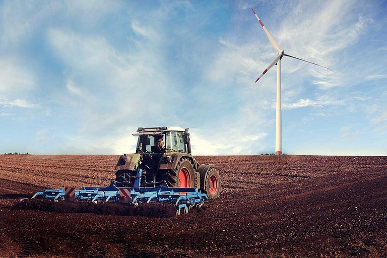 Ein Landwirt fährt mit einem Traktor und Pflug auf einem unbepflanzten Feld. Im Hintergrund steht ein Windrad